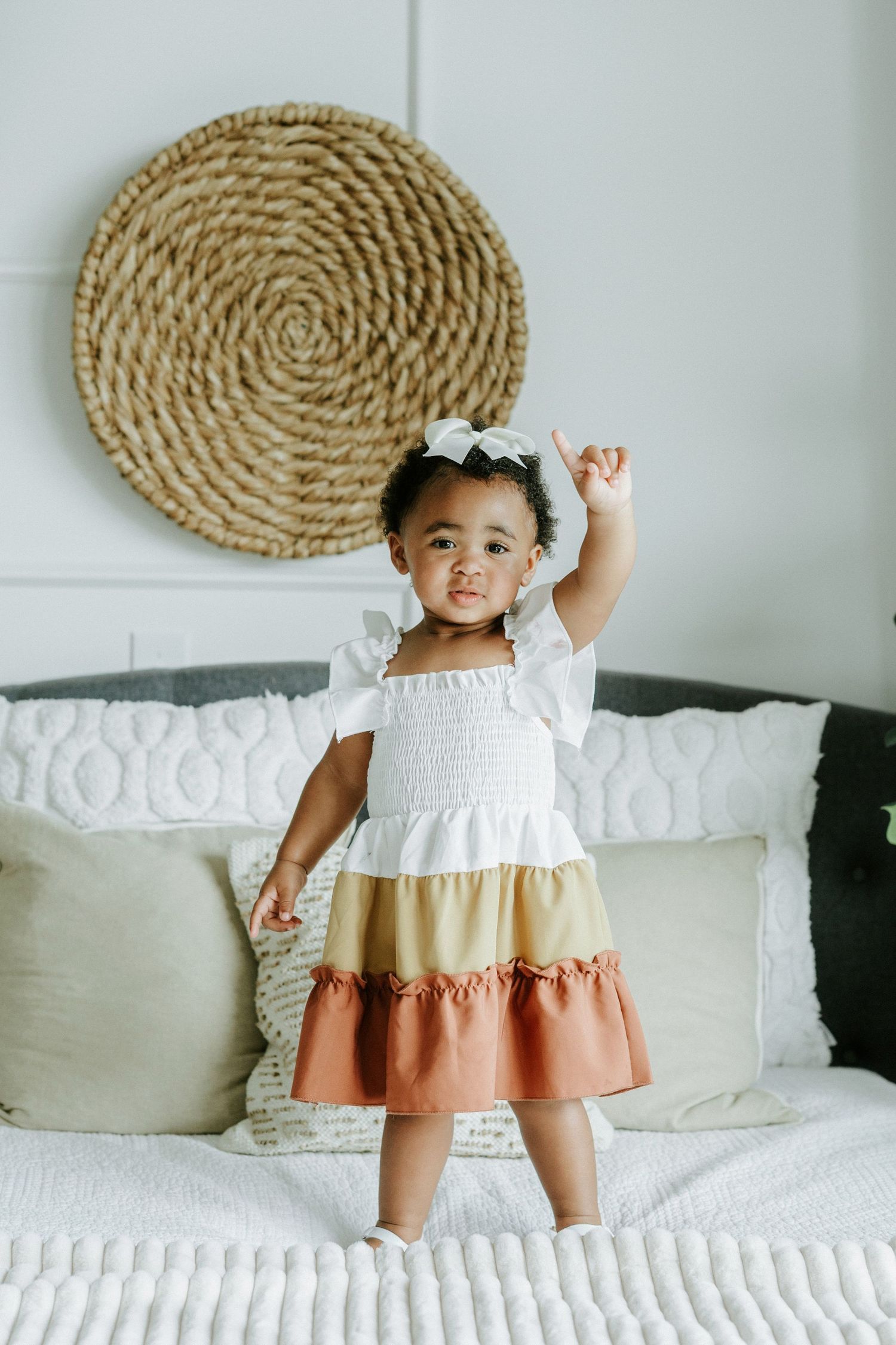 Toddler in tiered dress poses happily in front of woven wall decor on white bedding.