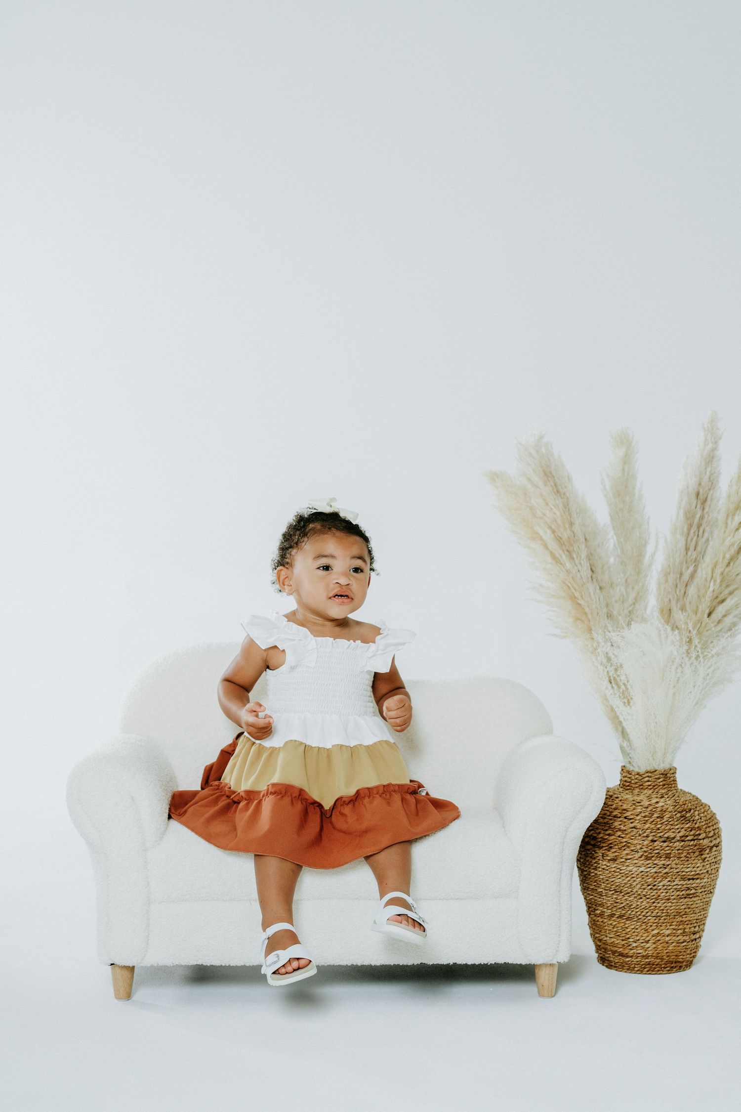 Small child in tricolor dress sits on white couch next to pampas grass decor.