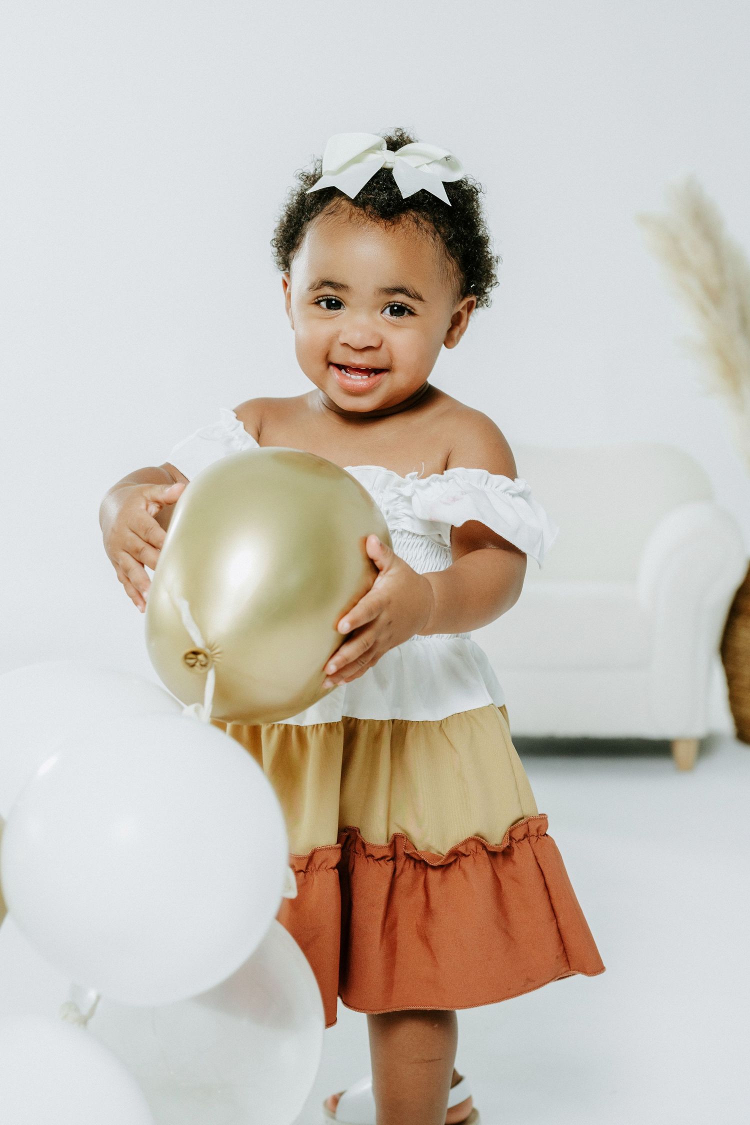 Happy toddler in white and tan dress holding gold balloon against white background.