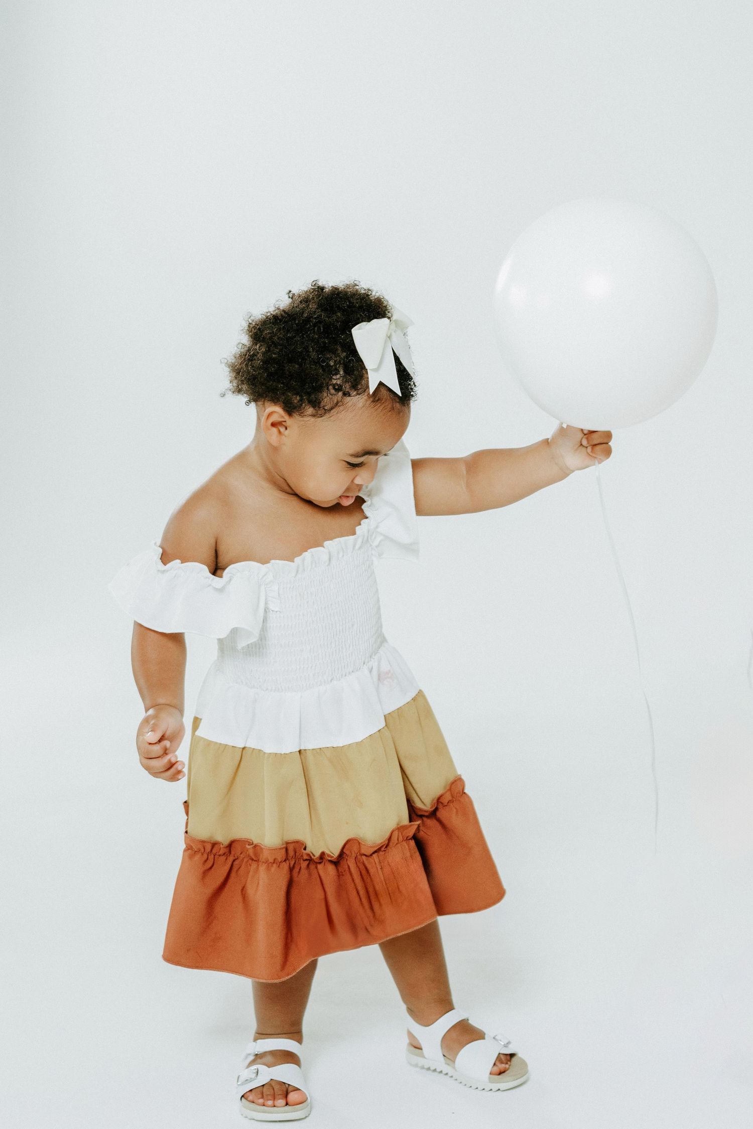 Toddler in tricolor dress plays with white balloon against minimal background.