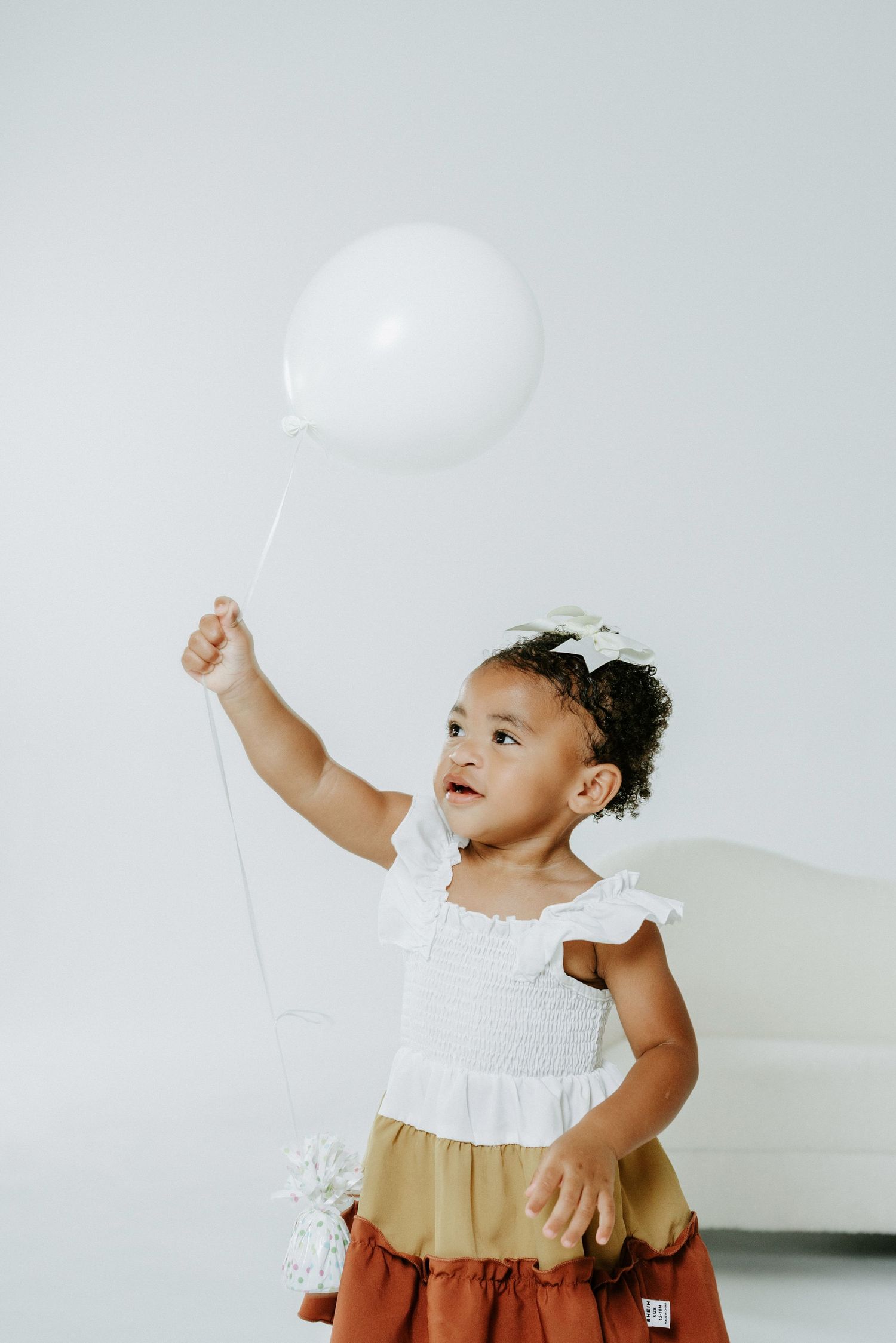 Small child holds white balloon while wearing tiered dress in studio setting.