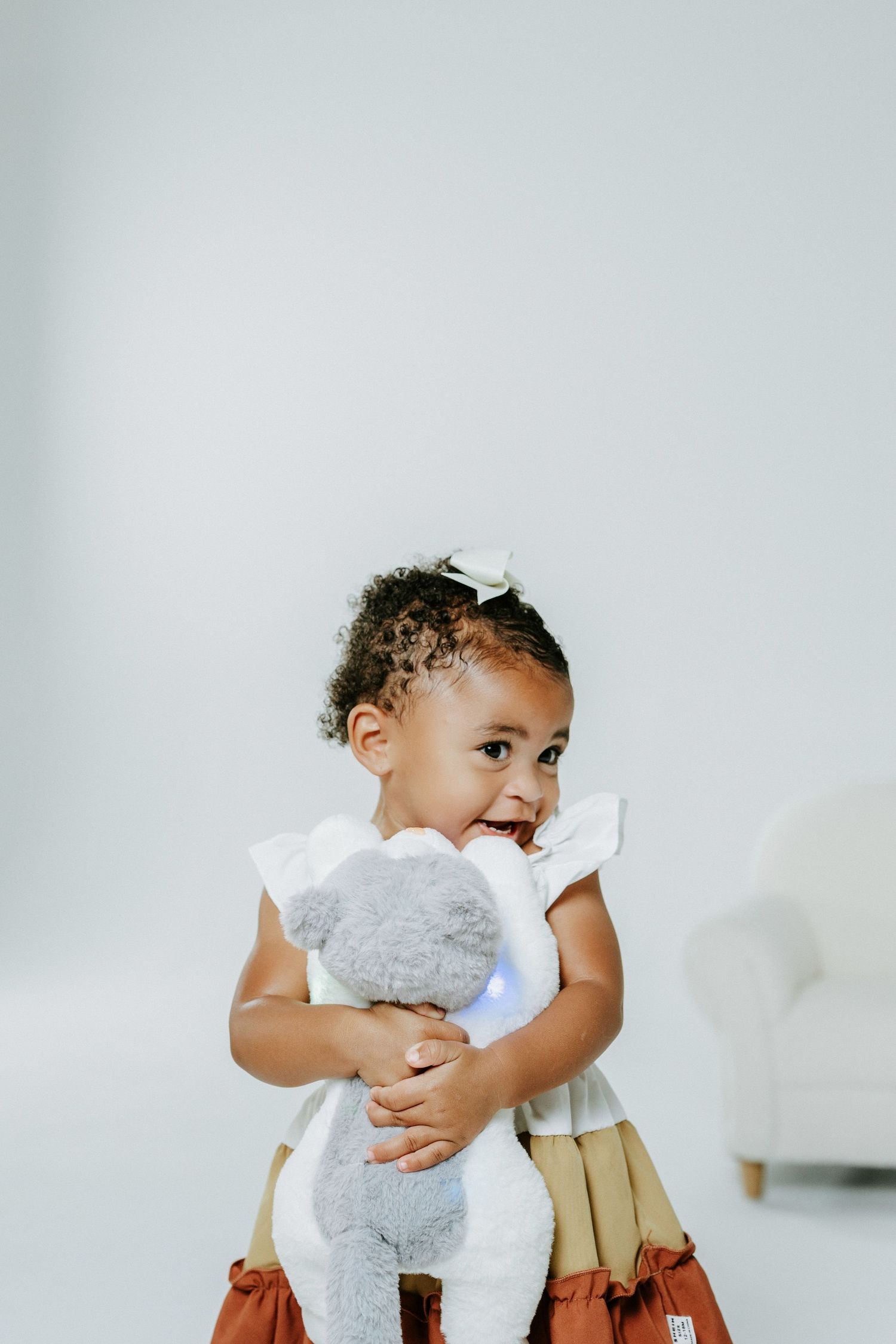 A small child holding a plush white teddy bear while wearing a white and tan tiered dress.