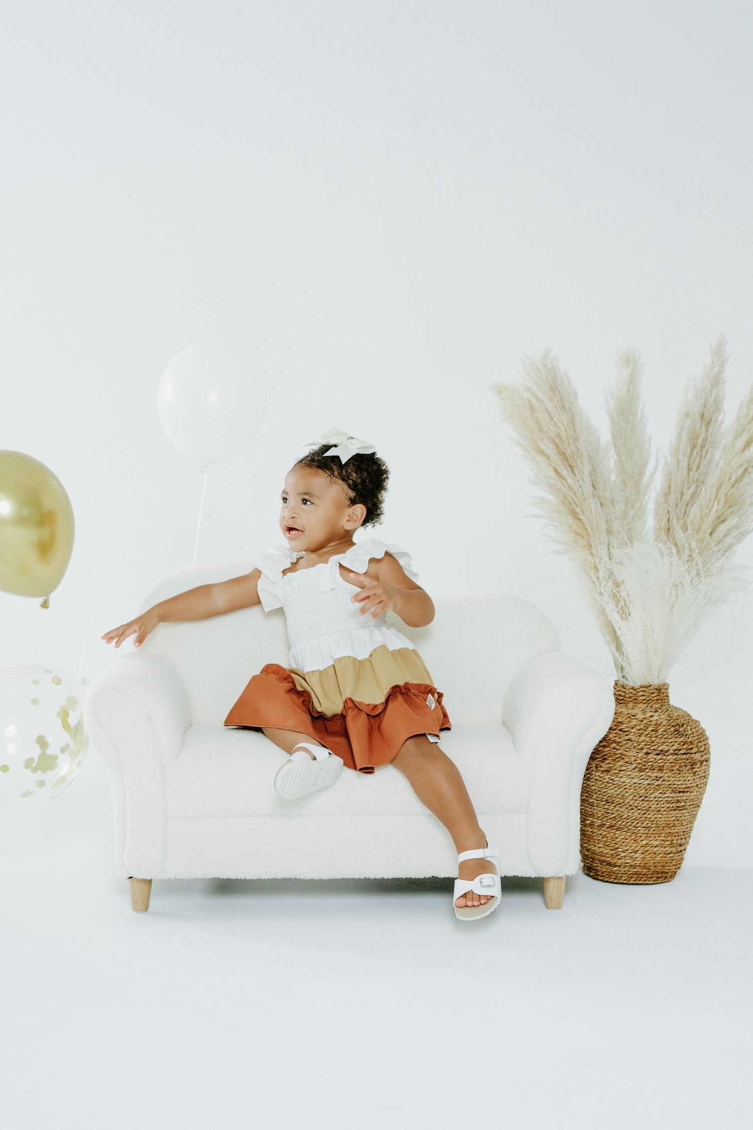 Playful toddler jumps on white couch wearing tricolor dress next to decorative vase.
