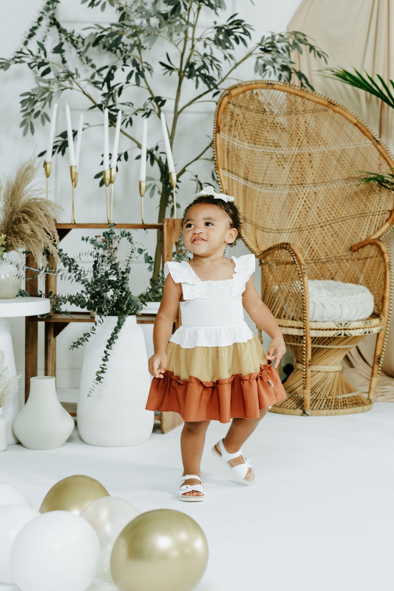 A toddler in a tricolor dress poses near a peacock chair and decorative plants.