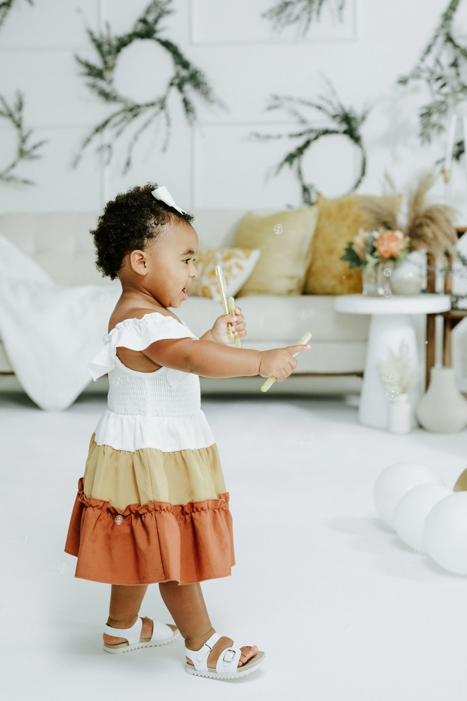 A toddler in a tiered dress holds party decorations in a minimalist white room setting.