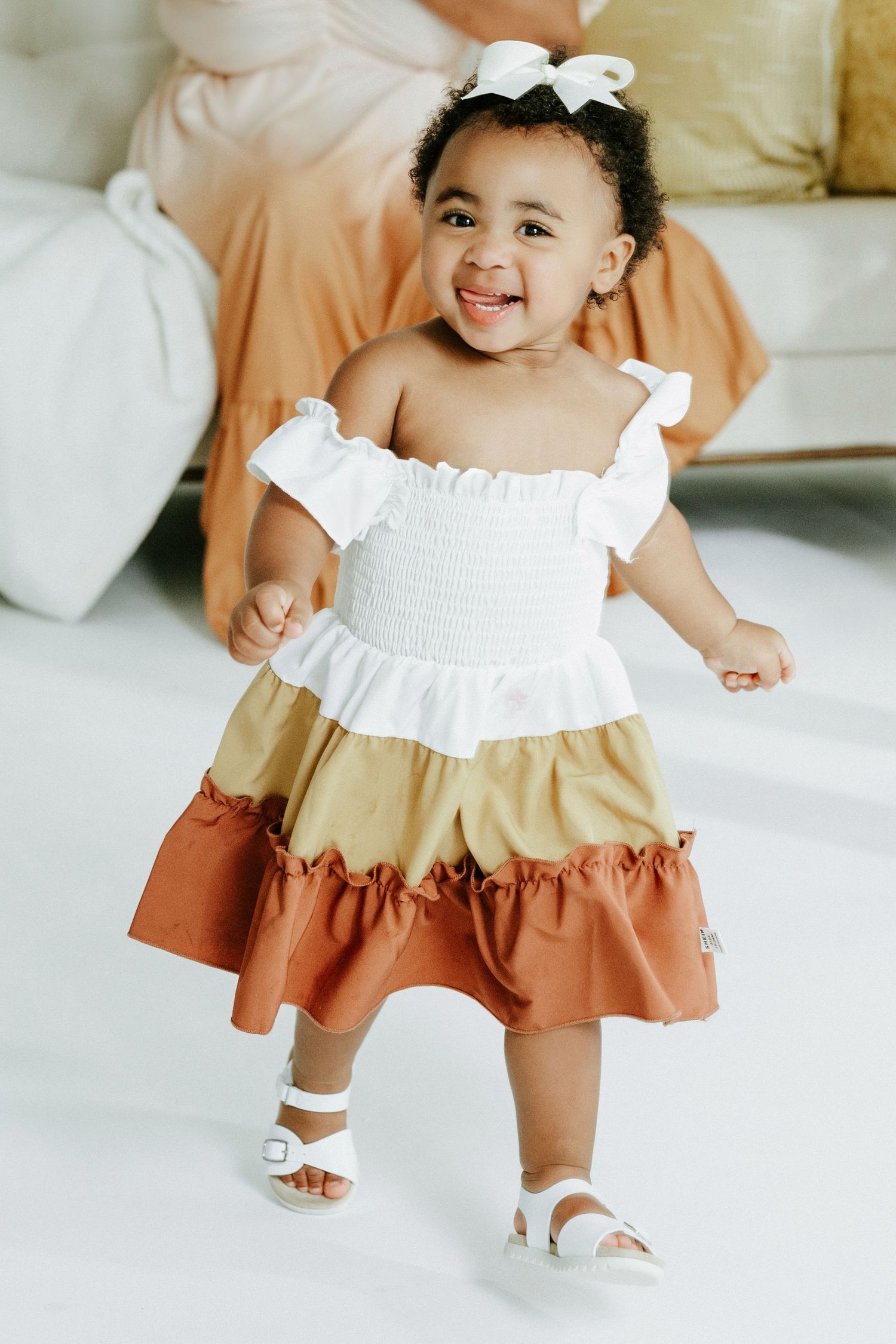 A toddler in a white, tan and rust colored dress smiles while standing in a bright studio setting.