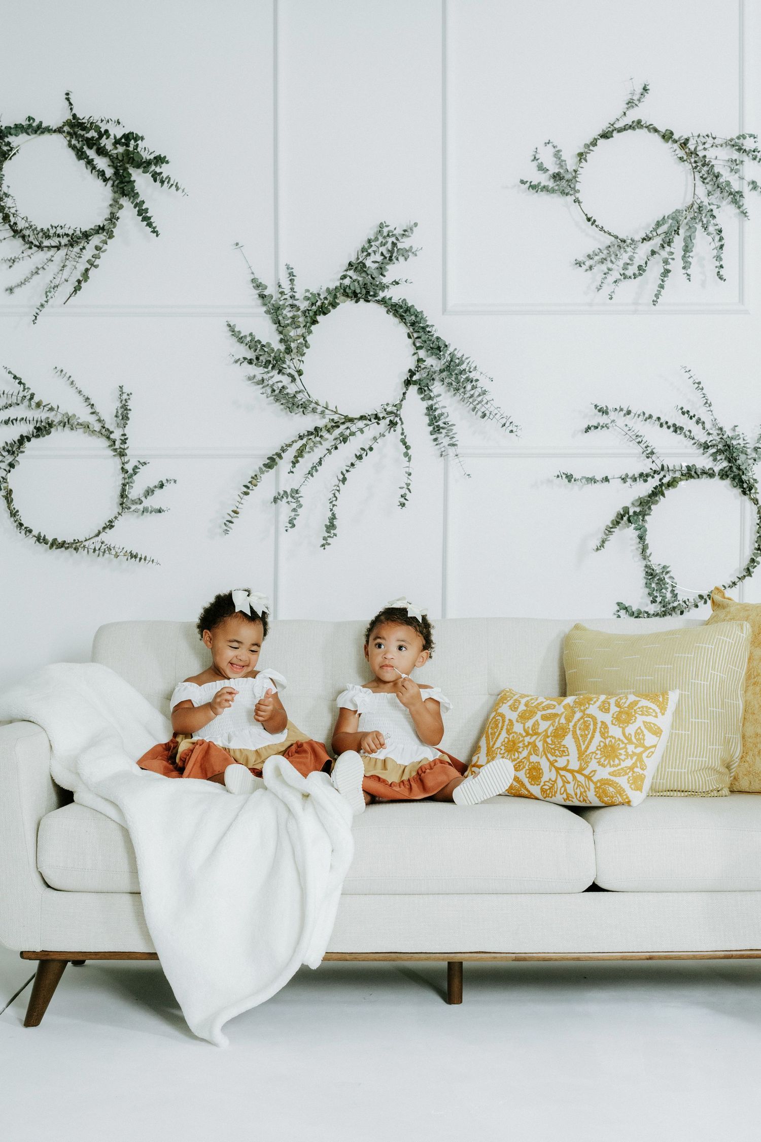 Two toddlers in matching outfits sit on a modern white couch beneath wall decorations.