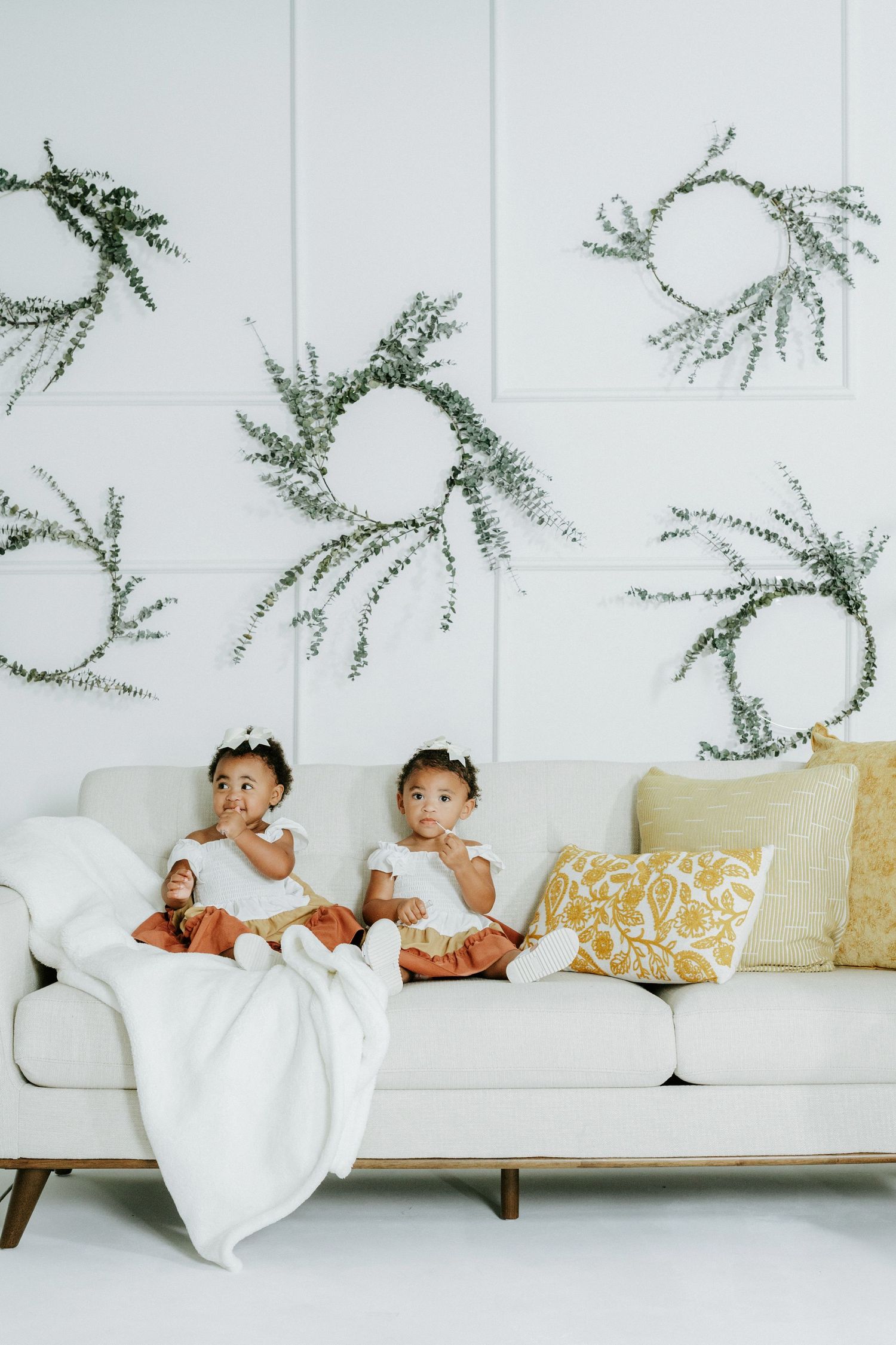 Two little ones sitting on a white couch under botanical wall decorations.