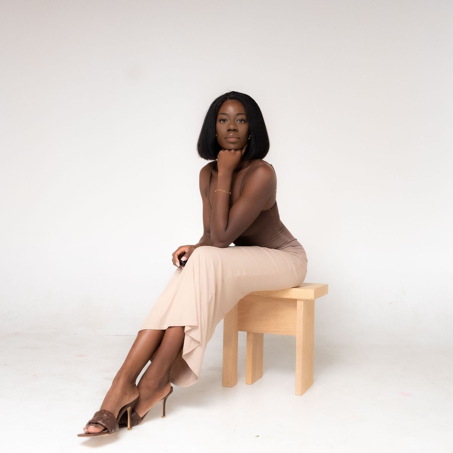 A woman in a beige dress sits gracefully on a wooden stool against a white background while posing elegantly.
