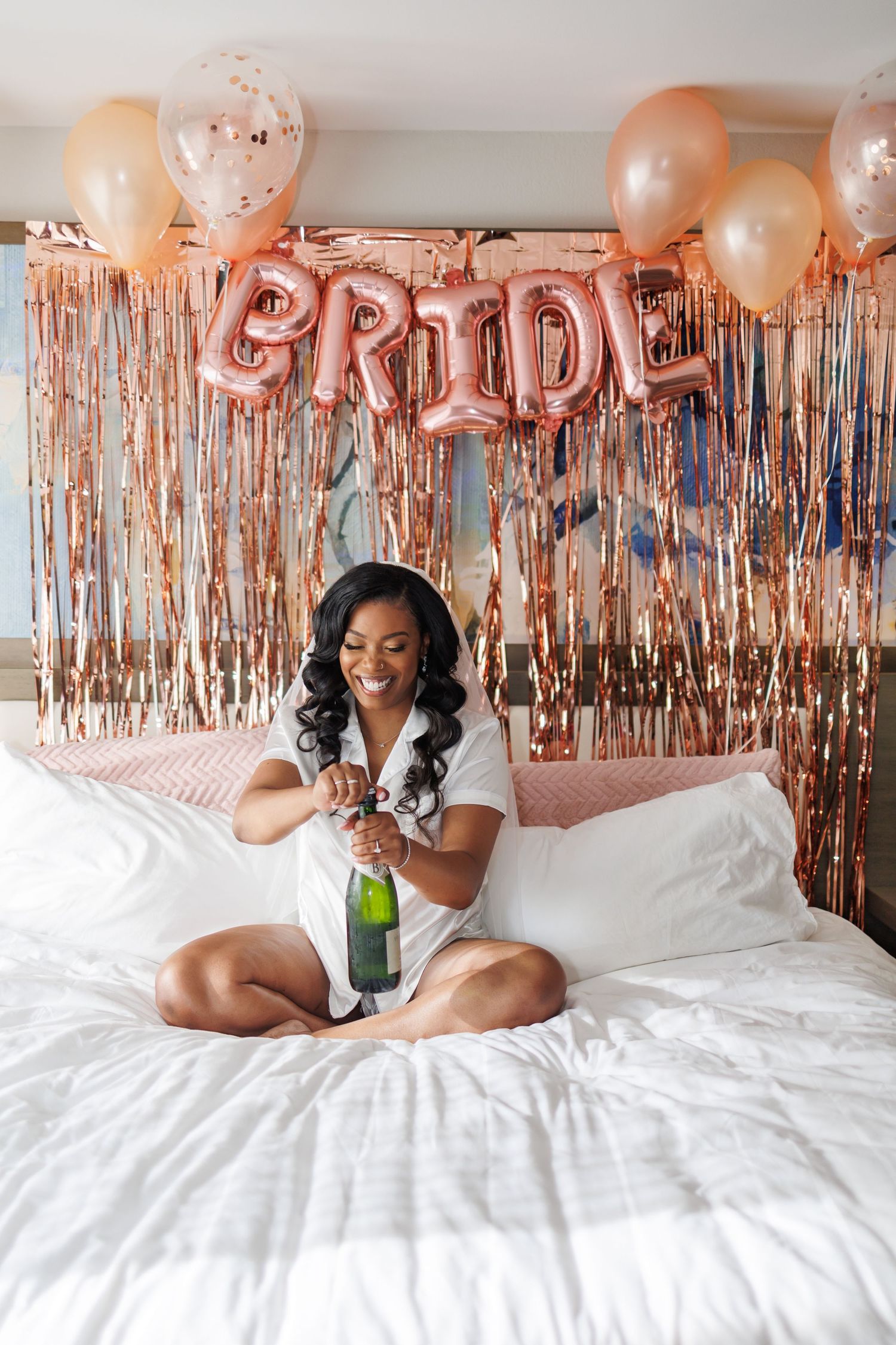 A bride in white pajamas popping champagne as she gets ready during her wedding morning in a hotel room decorated in rose gold