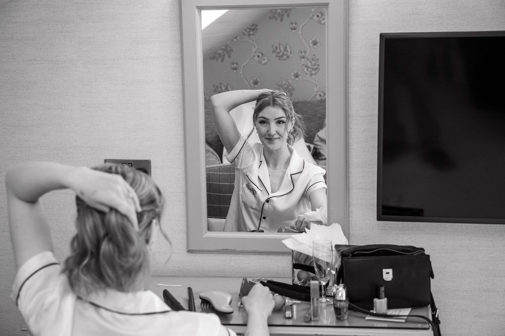 Bride getting ready, styling her hair while looking in a mirror in black and white photo.
