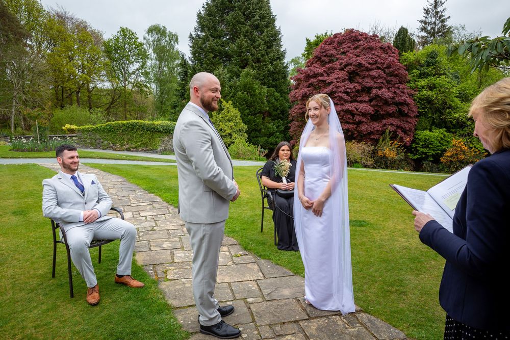 Outdoor wedding ceremony on a garden path with groom in grey suit and bride in white dress with veil.