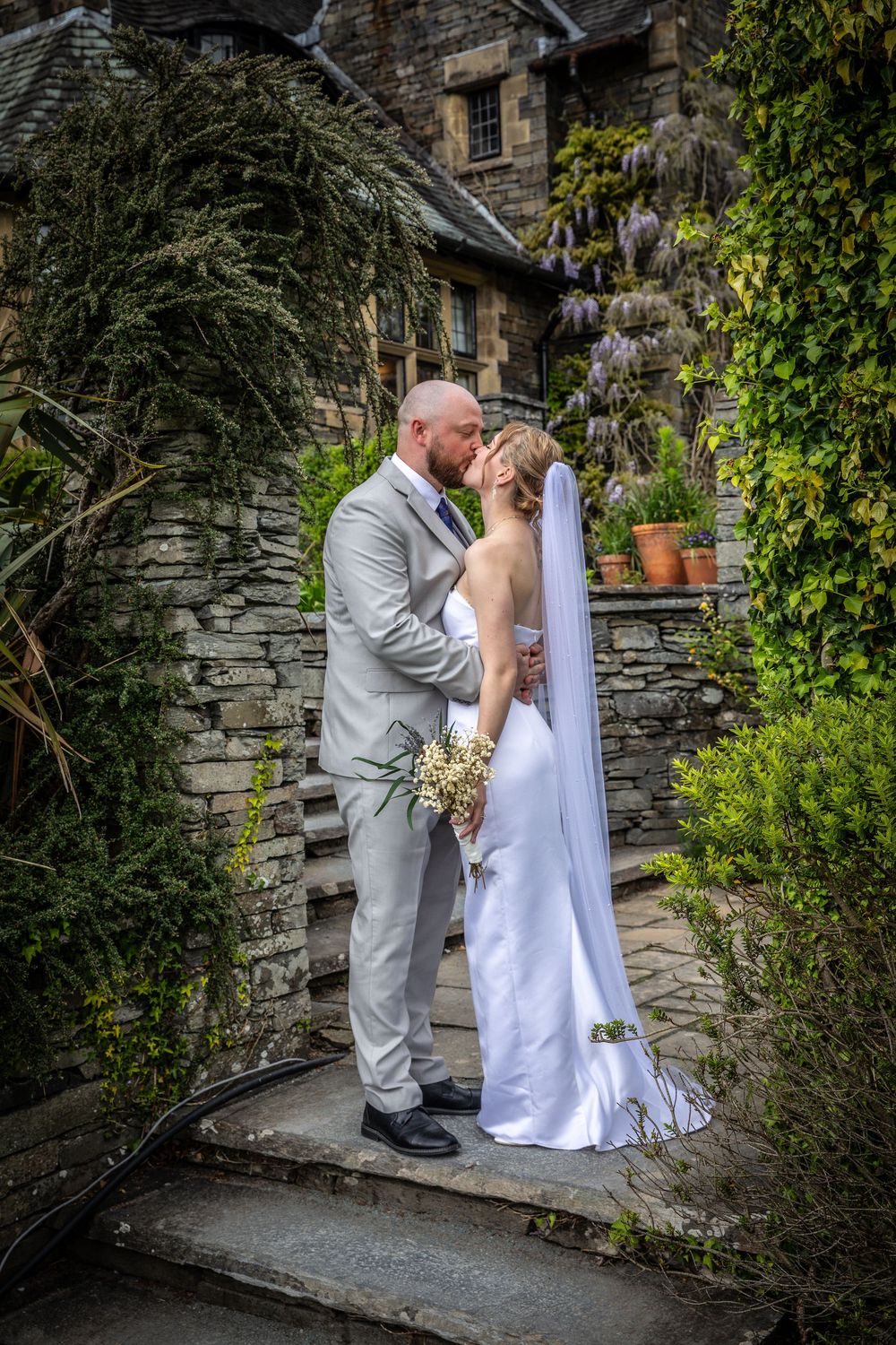 Bride and groom share a kiss on stone steps beside an ivy-covered stone building with lush greenery.