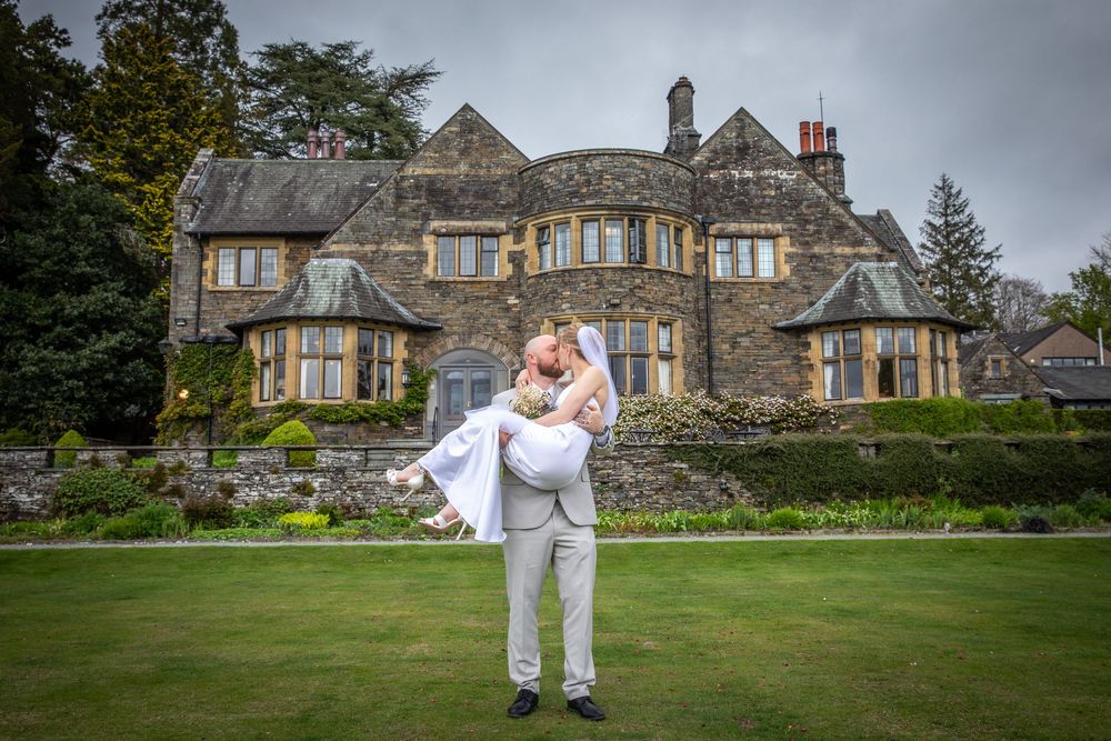 Groom lifts bride in white dress on lawn in front of a grand stone manor house on their wedding day.