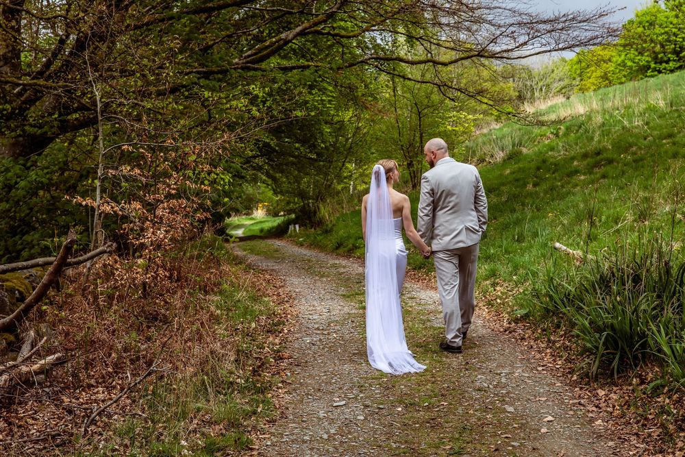 Bride in white dress and groom in grey suit walk hand in hand along a tree-lined woodland path.