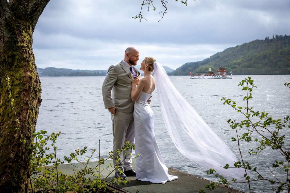 Bride and groom share a romantic moment by a lake with a veil flowing in the wind and hills beyond.