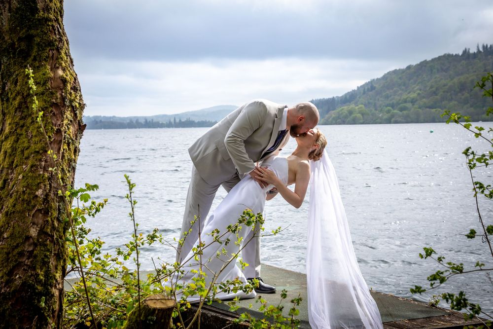 Bride and groom share a romantic dip kiss by a lakeside dock surrounded by lush greenery.