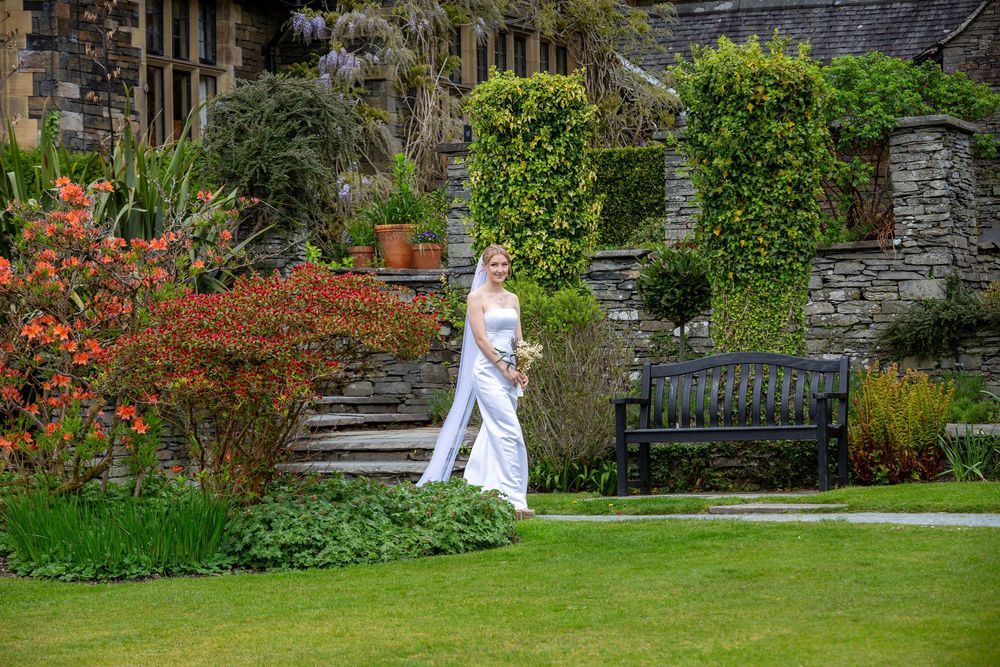 Bride in white gown walks through a beautiful garden with colorful flowers beside a stone manor house.