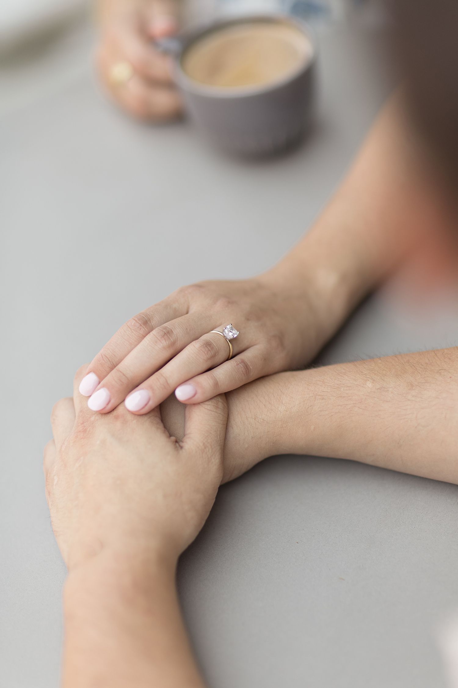 Close-up of hands holding each other with an engagement ring visible and a coffee cup in the background.