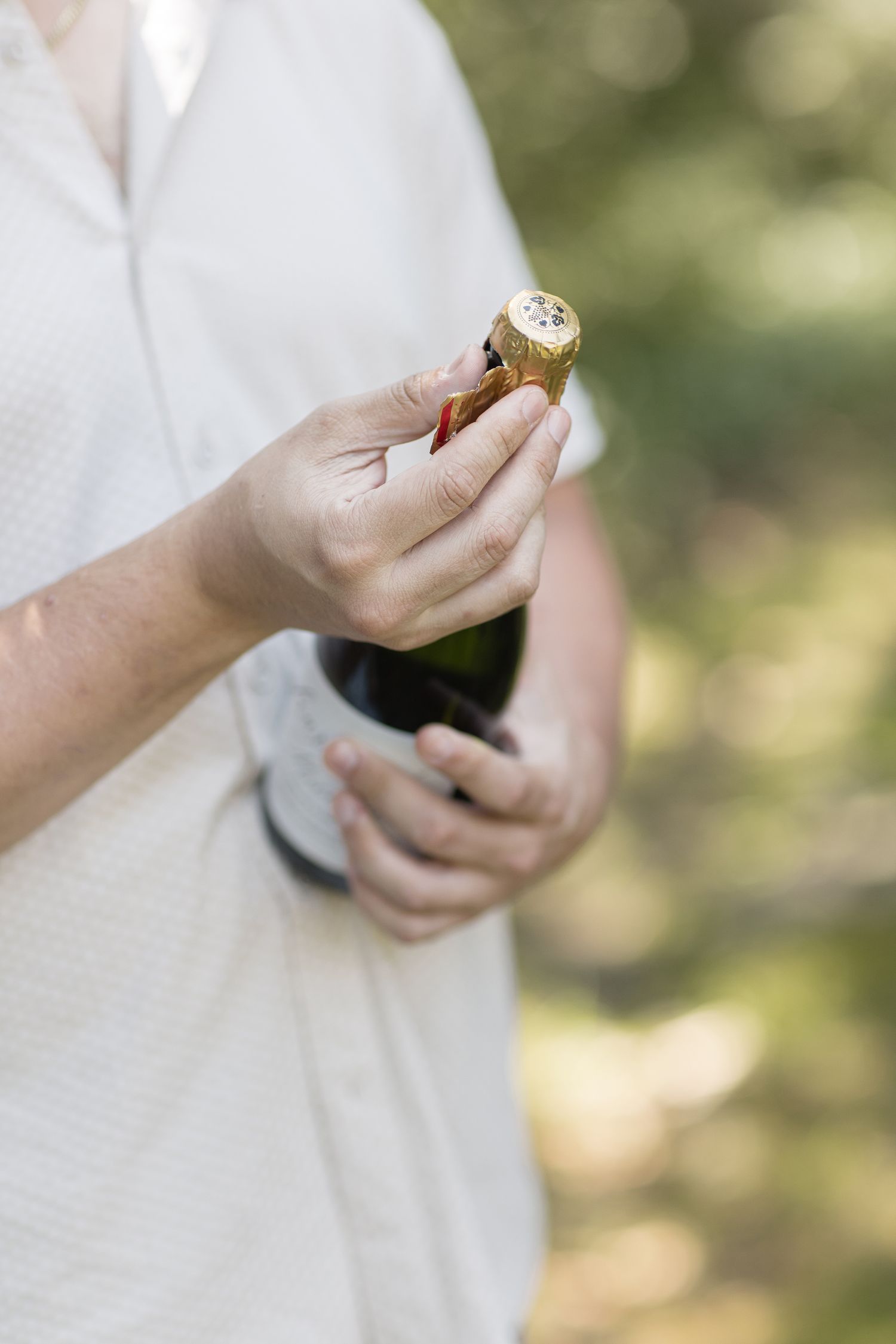 A couple shares a romantic moment outdoors while one holds a wine bottle among sunlit trees in the background.