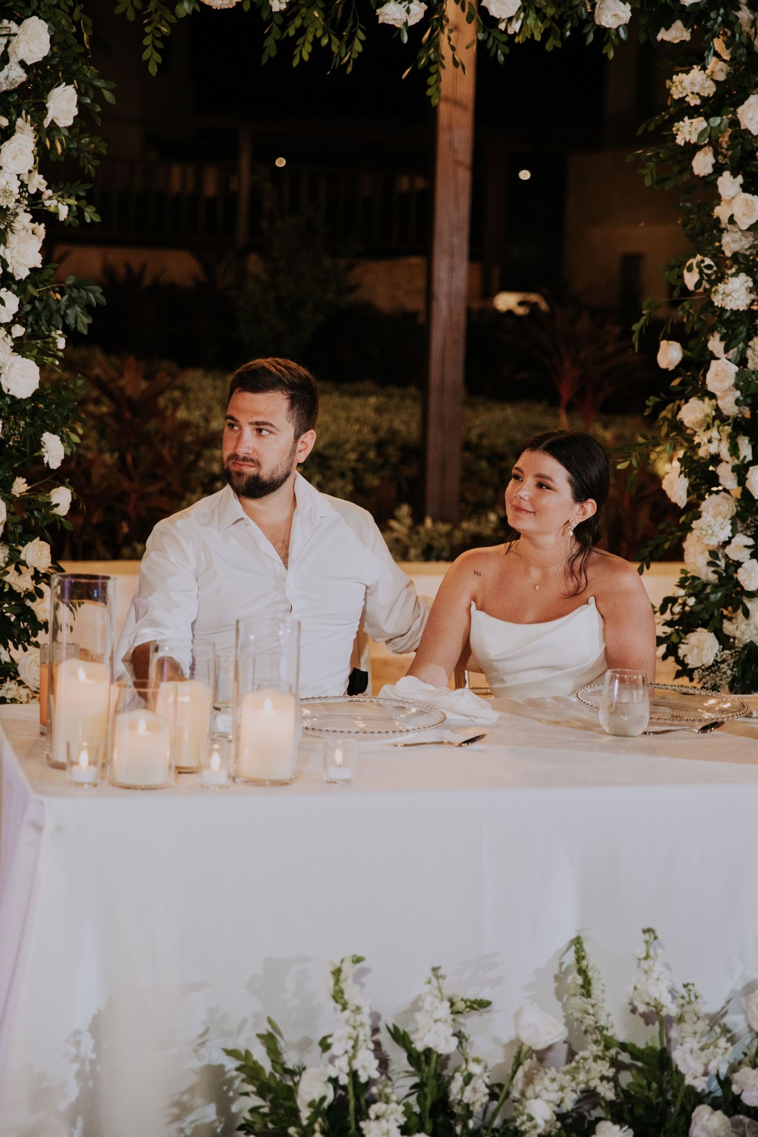 A couple sits at an elegant wedding reception table decorated with candles and floral arrangements.