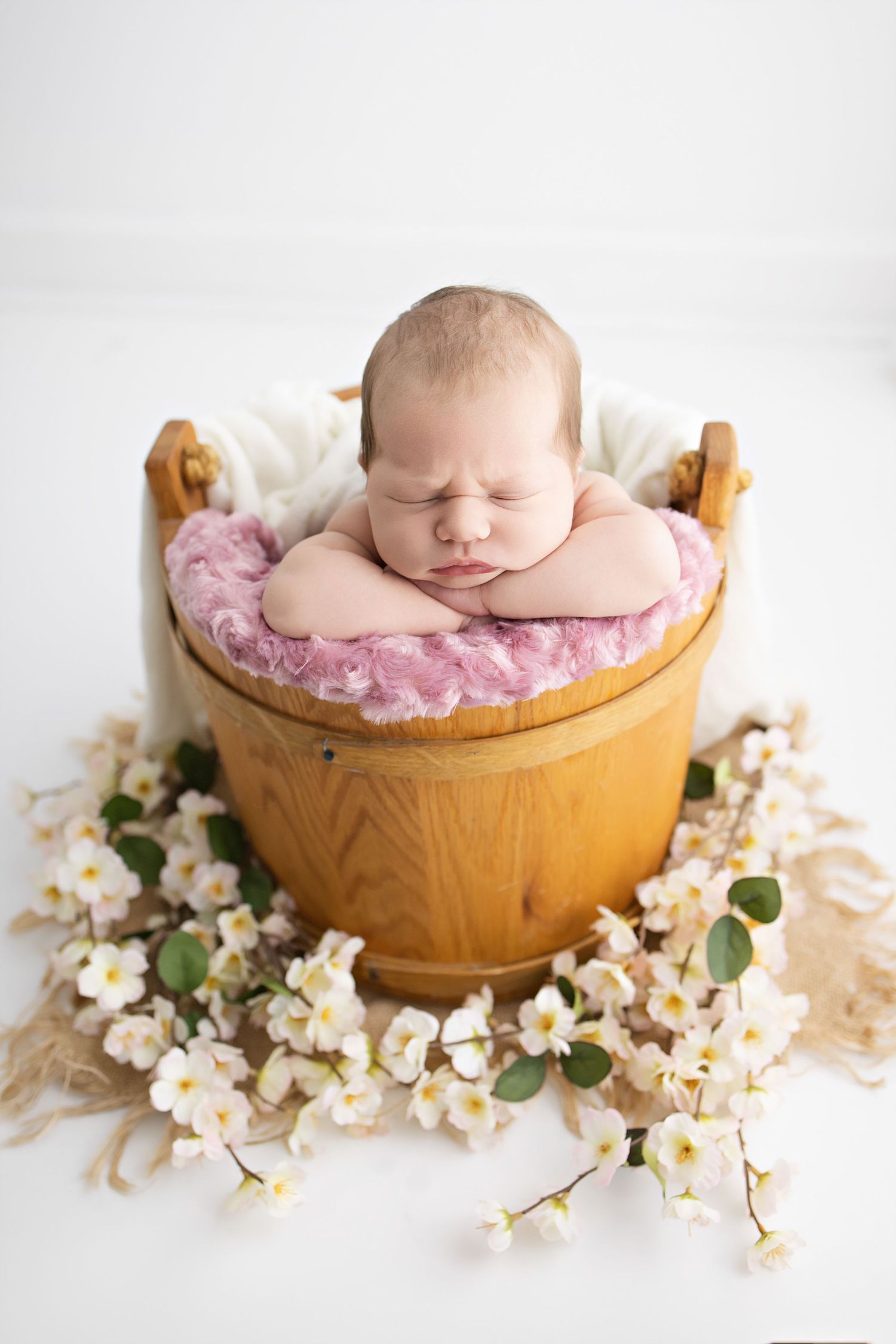 Sleeping newborn in wooden bucket prop with pink cushioning surrounded by delicate white flower blossoms.