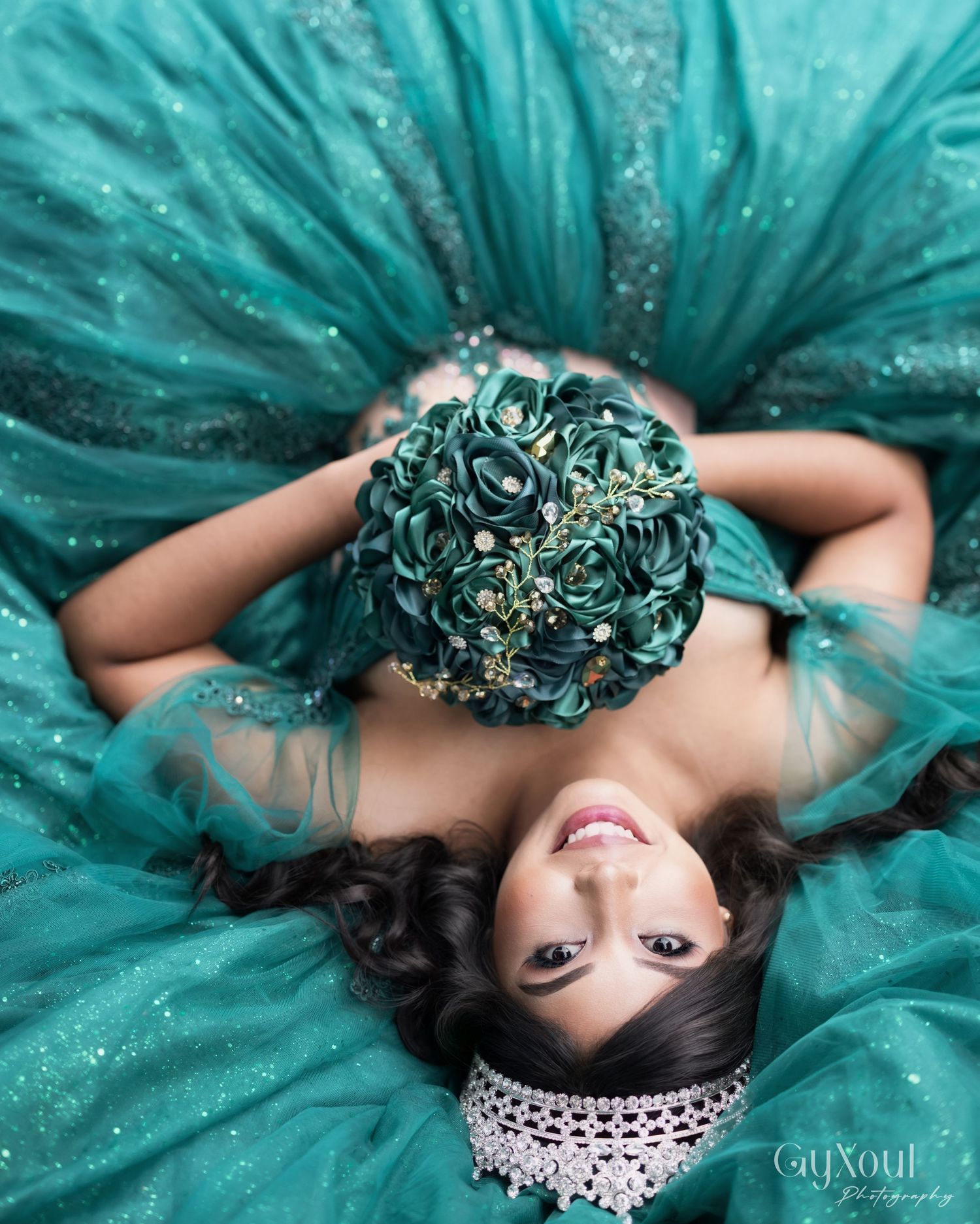A glamorous turquoise gown with glitter sparkles fills the frame as a woman lies holding a matching floral bouquet.