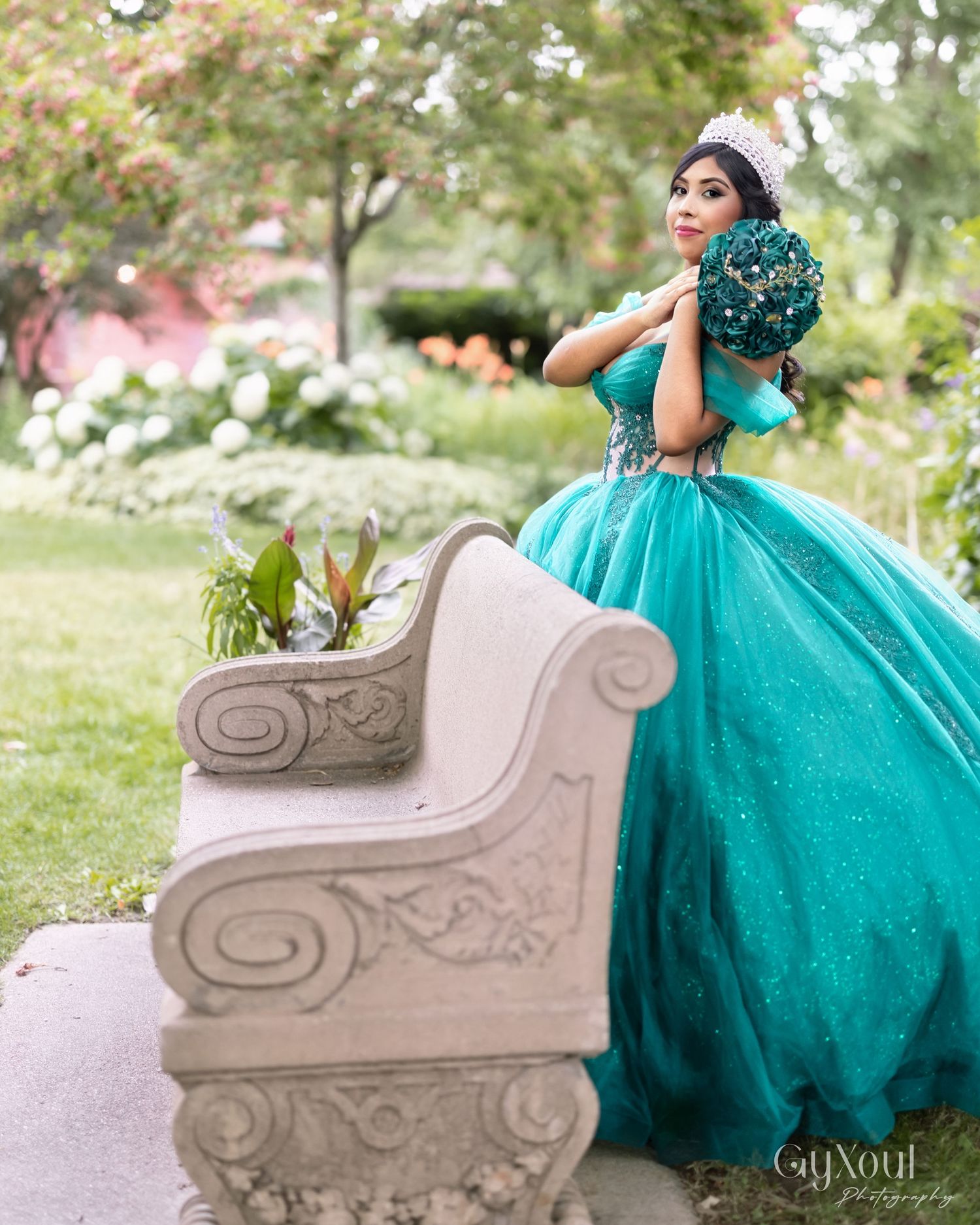 A stunning turquoise quinceañera ball gown photographed in a garden setting with an ornate stone bench.