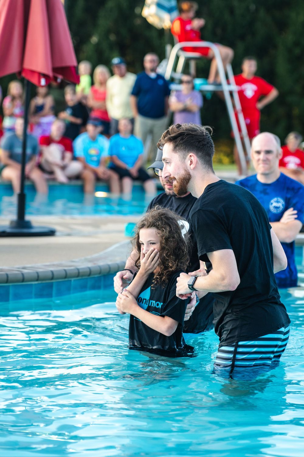 People participate in a baptism ceremony at an outdoor swimming pool with onlookers watching from poolside.