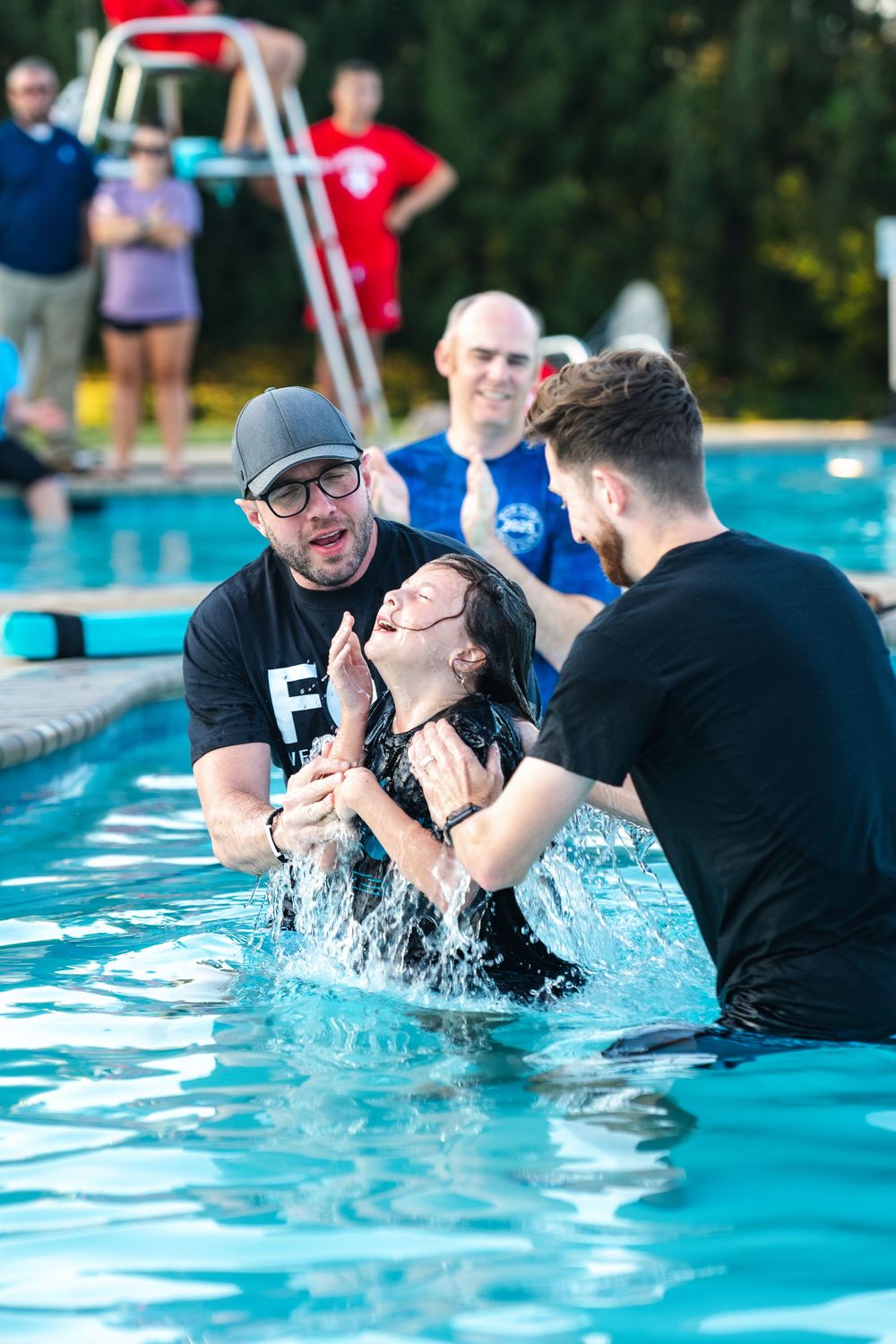 People participate in a baptism ceremony in an outdoor swimming pool surrounded by trees on a sunny day.