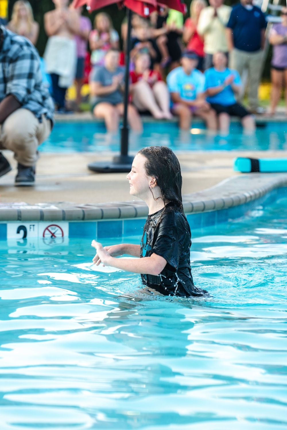 Someone in swimming attire enjoys a sunny day at a community pool with onlookers visible in the background.