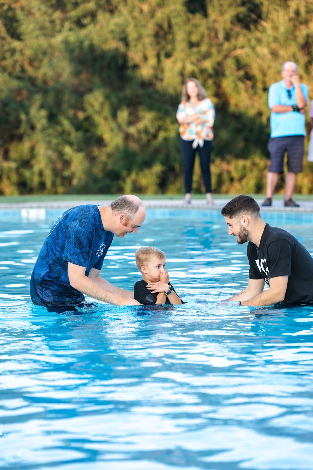 A baptism ceremony takes place in a bright blue swimming pool on a sunny day.