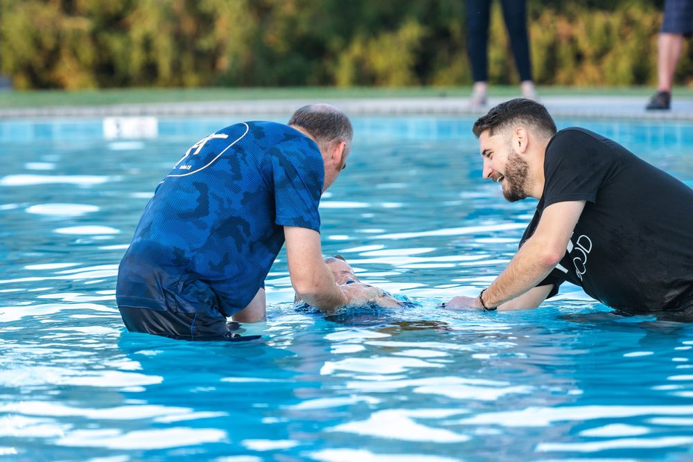 A baptism ceremony takes place in an outdoor swimming pool on a sunny day.