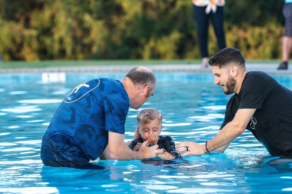 Two people in blue and black help a child in bright blue water during a baptism ceremony at an outdoor pool.