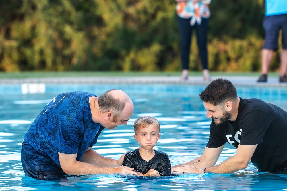 A baptism ceremony takes place in a bright blue swimming pool on a sunny day.