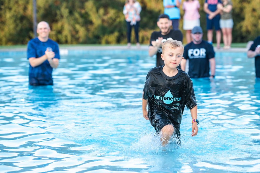 A baptism ceremony takes place in a bright blue swimming pool on a sunny day.