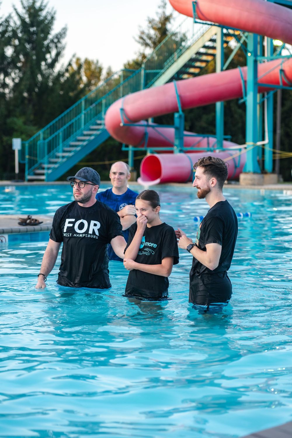 A baptism ceremony takes place in a bright blue swimming pool on a sunny day.