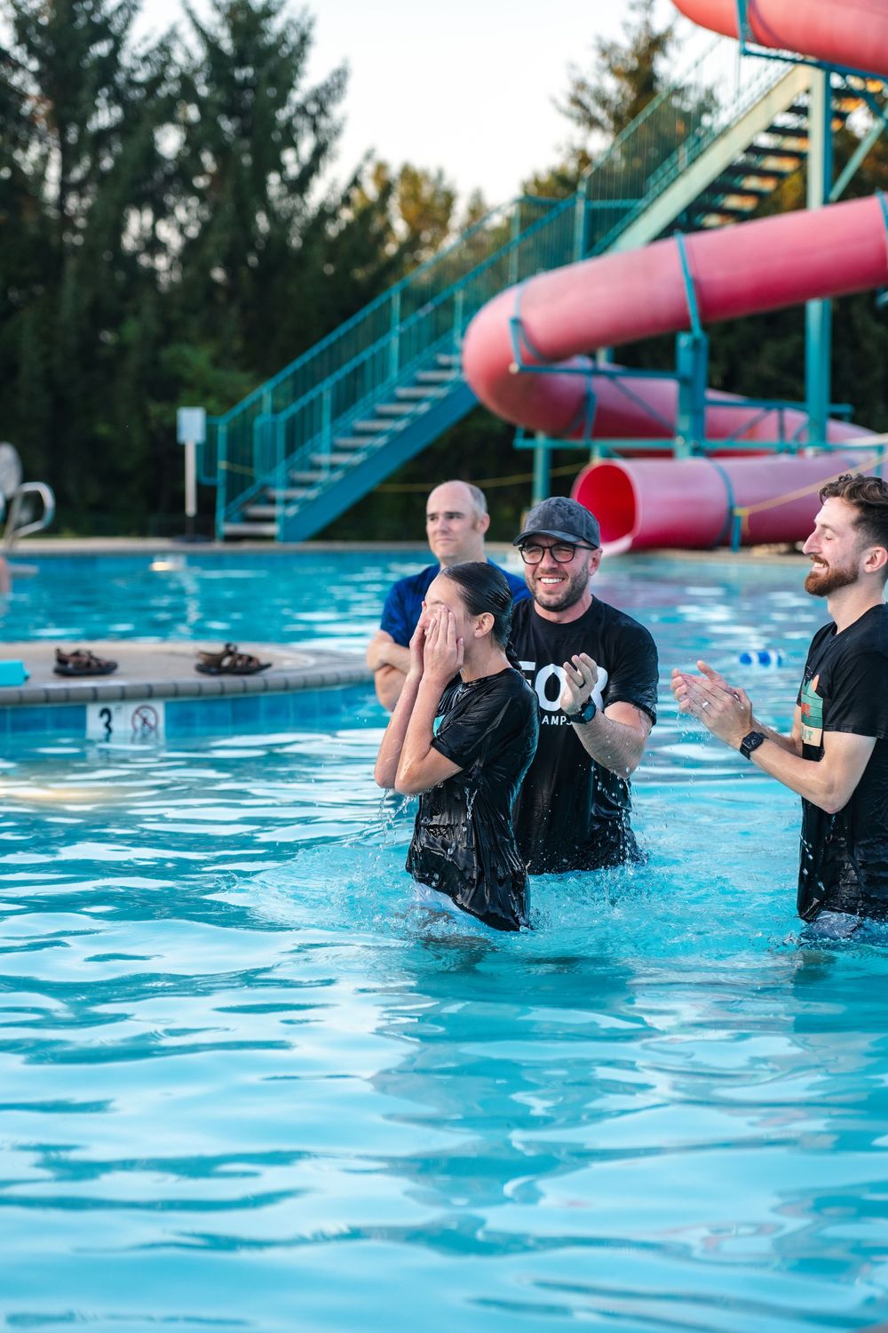 A baptism ceremony takes place in a bright blue swimming pool on a sunny day.