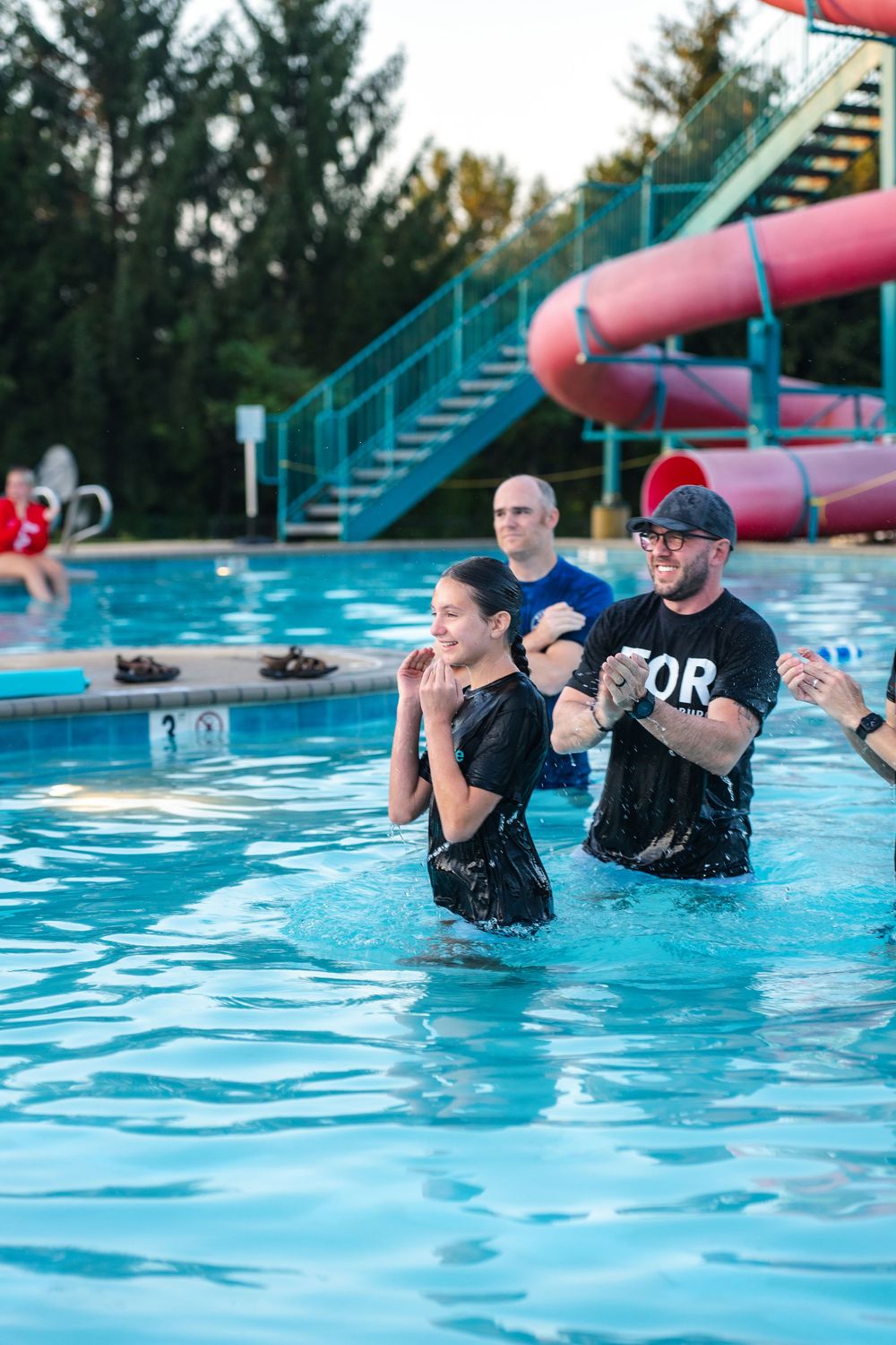 A baptism ceremony takes place in a bright blue swimming pool on a sunny day.