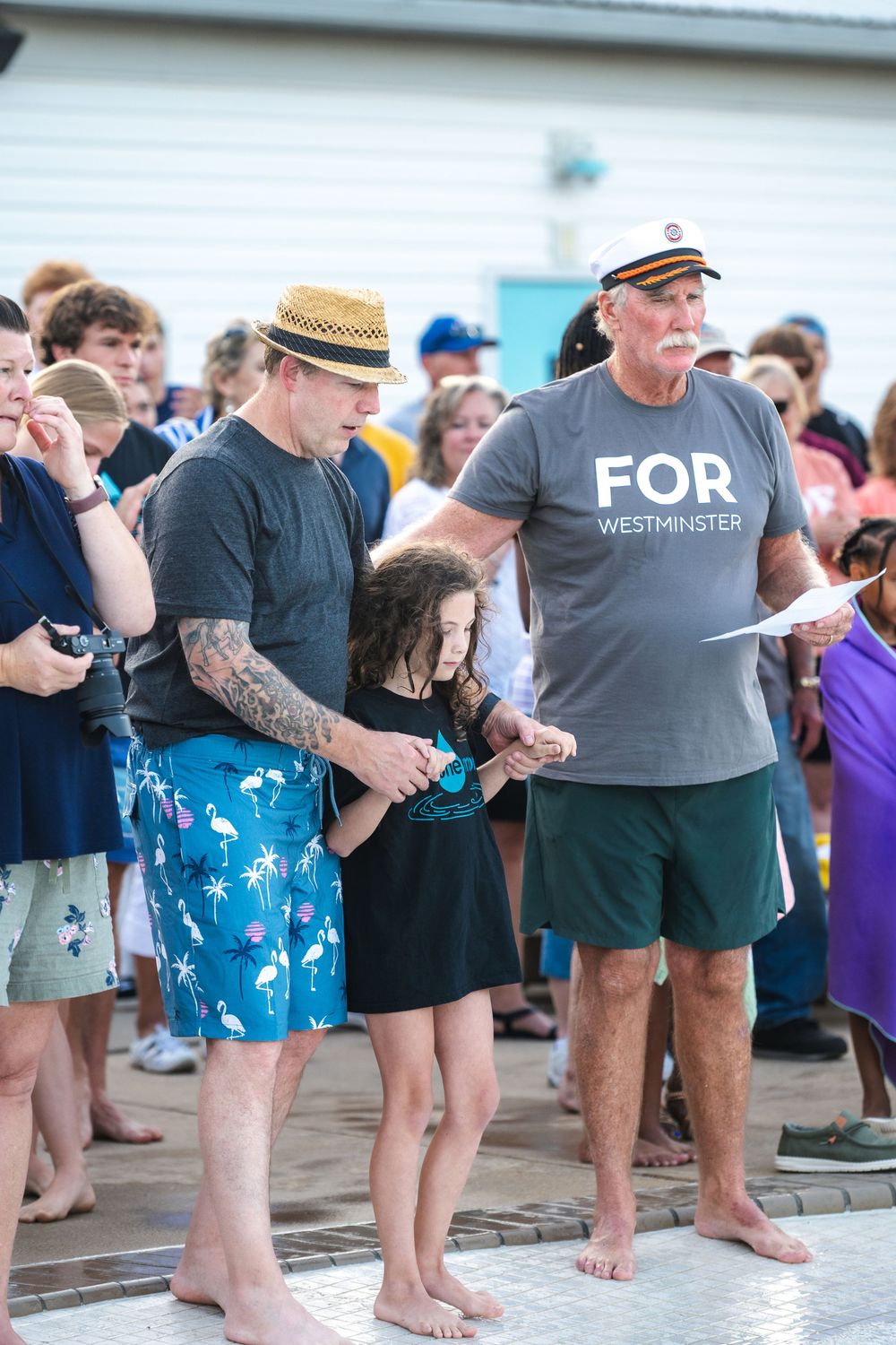 People gather barefoot at an outdoor baptism with someone wearing a nautical captain's hat and 'FOR' t-shirt.
