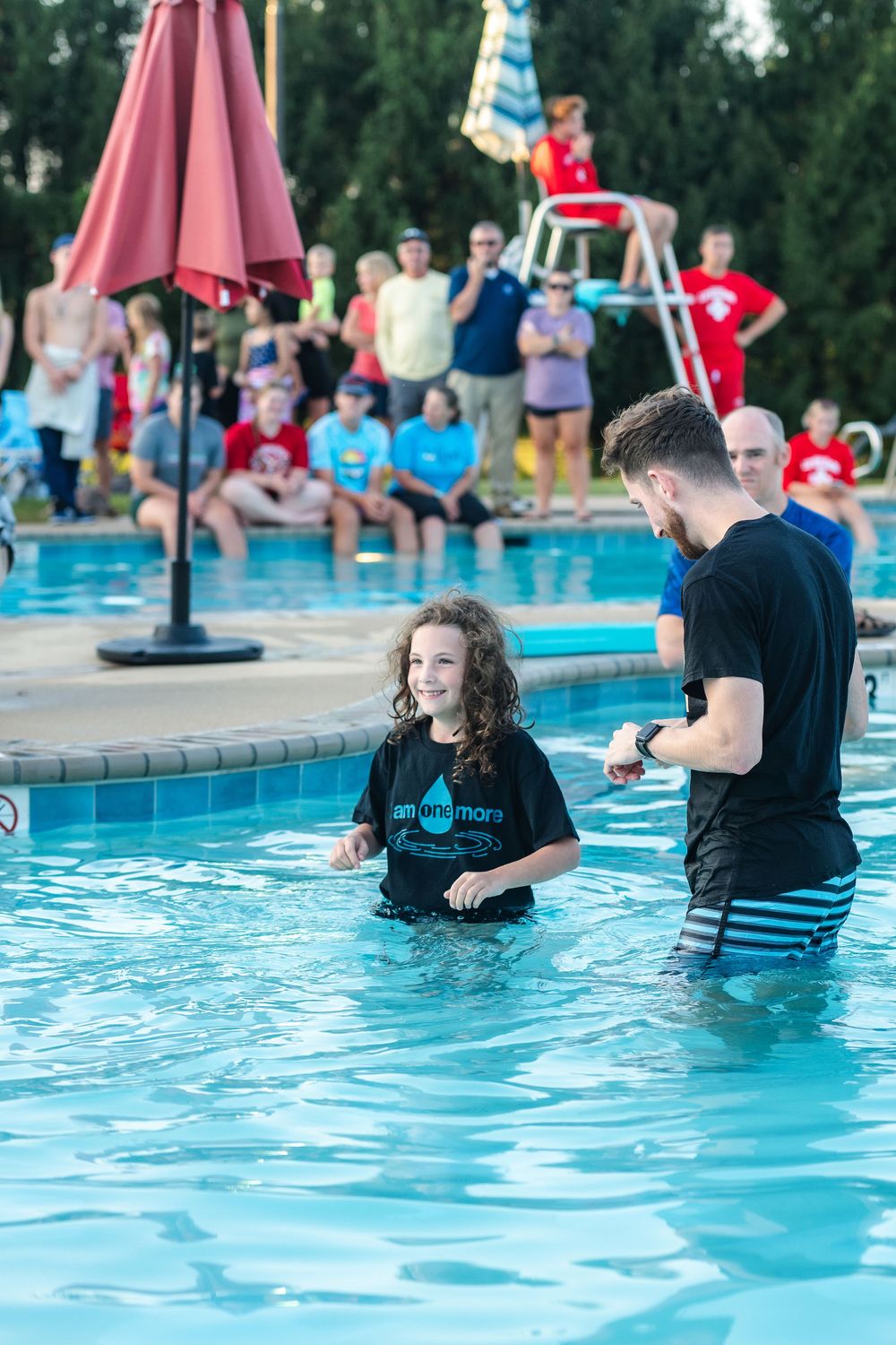 An outdoor baptism ceremony takes place in a swimming pool while onlookers watch from the pool deck area.