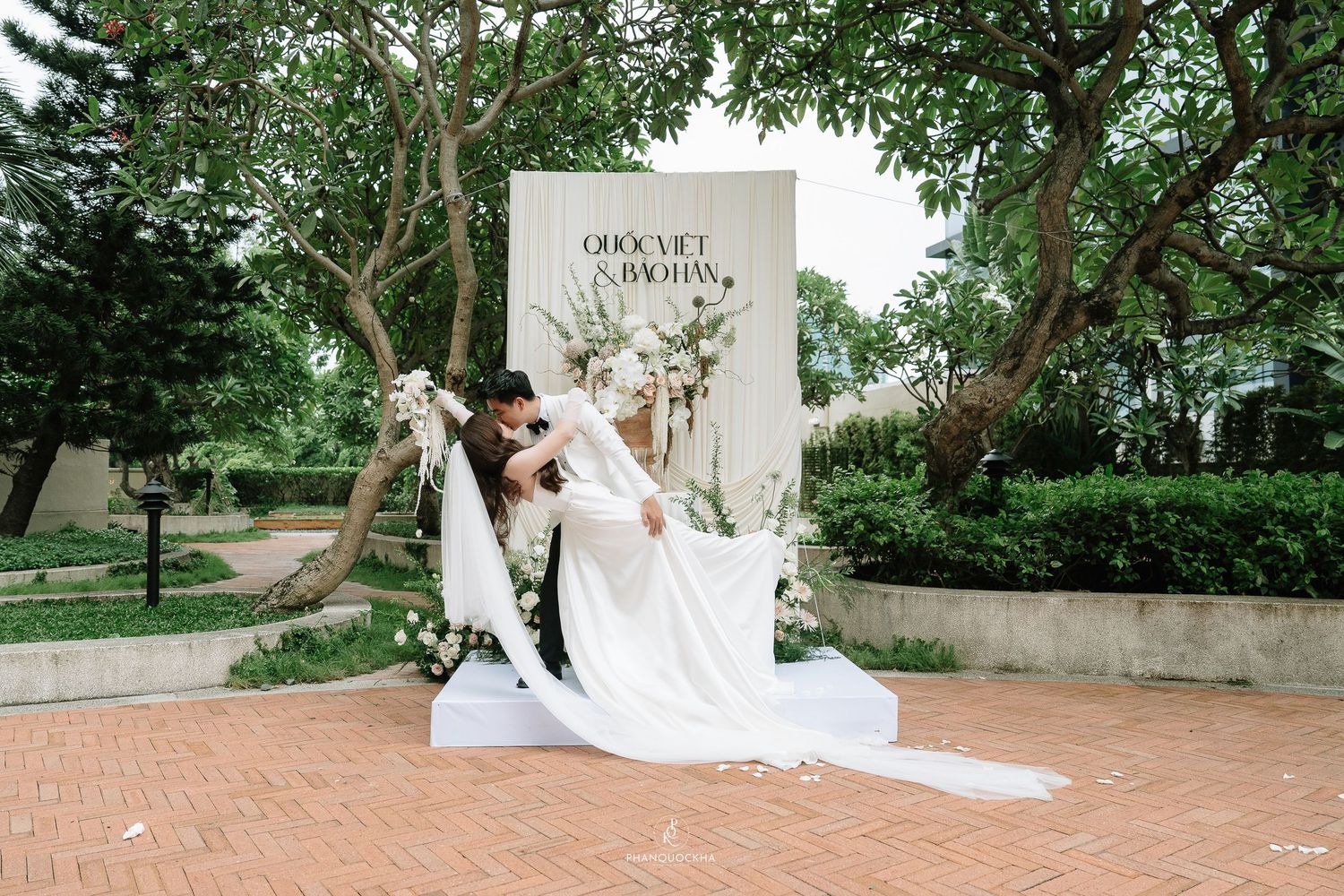 Bride and groom share romantic kiss on white platform stage before elegant floral wedding backdrop in lush garden setting.