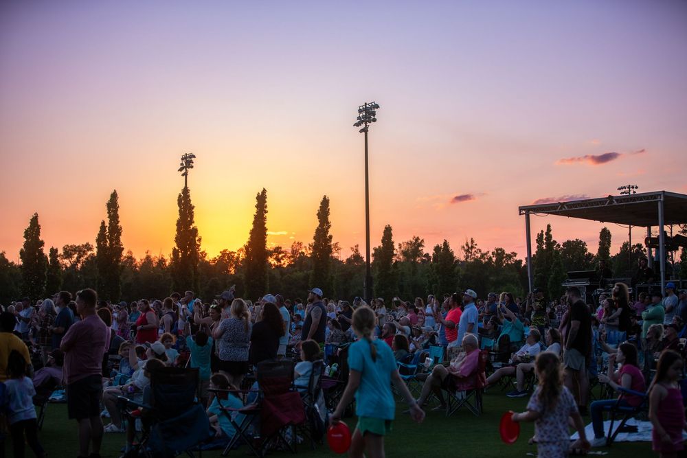 Large crowd enjoys an outdoor concert at sunset, with warm orange and purple skies illuminating a festive summer evening event.