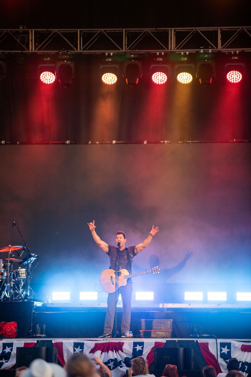 A guitarist performs on a patriotic-themed stage with dramatic red and blue concert lighting and raised arms.
