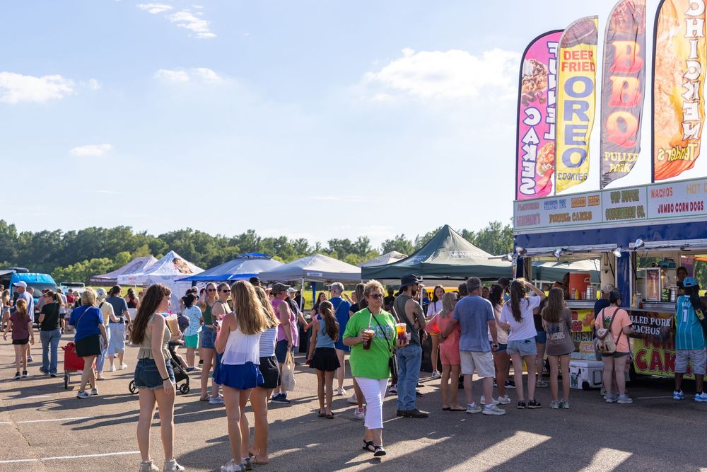 Crowds browse food vendor booths at an outdoor fair on a sunny day, with Oreo and taco signs visible.
