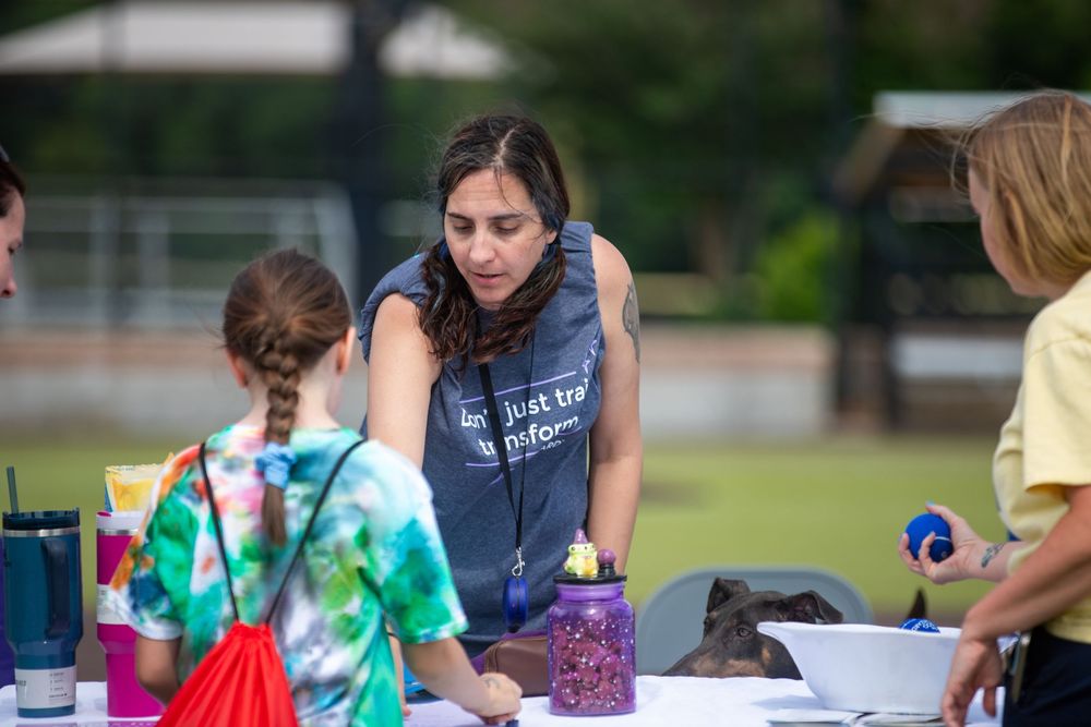 Woman in gray tank top assists children at outdoor craft table during school or community event.