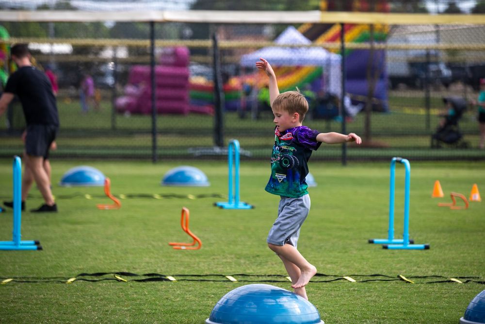 Young boy balancing on blue dome obstacle during outdoor kids sports activity day with colorful cones and hurdles.
