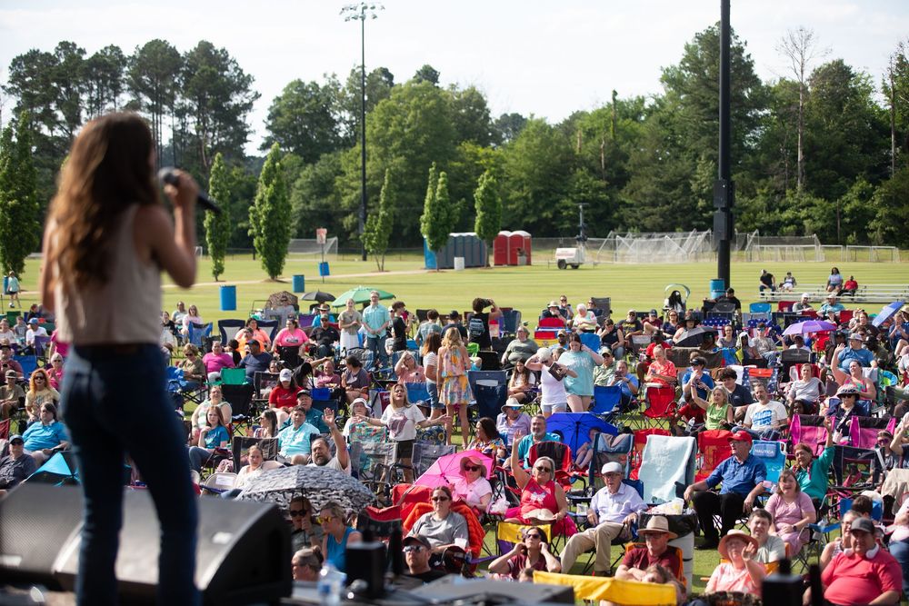 Outdoor concert with a female performer on stage entertaining a large crowd of families seated on lawn chairs.
