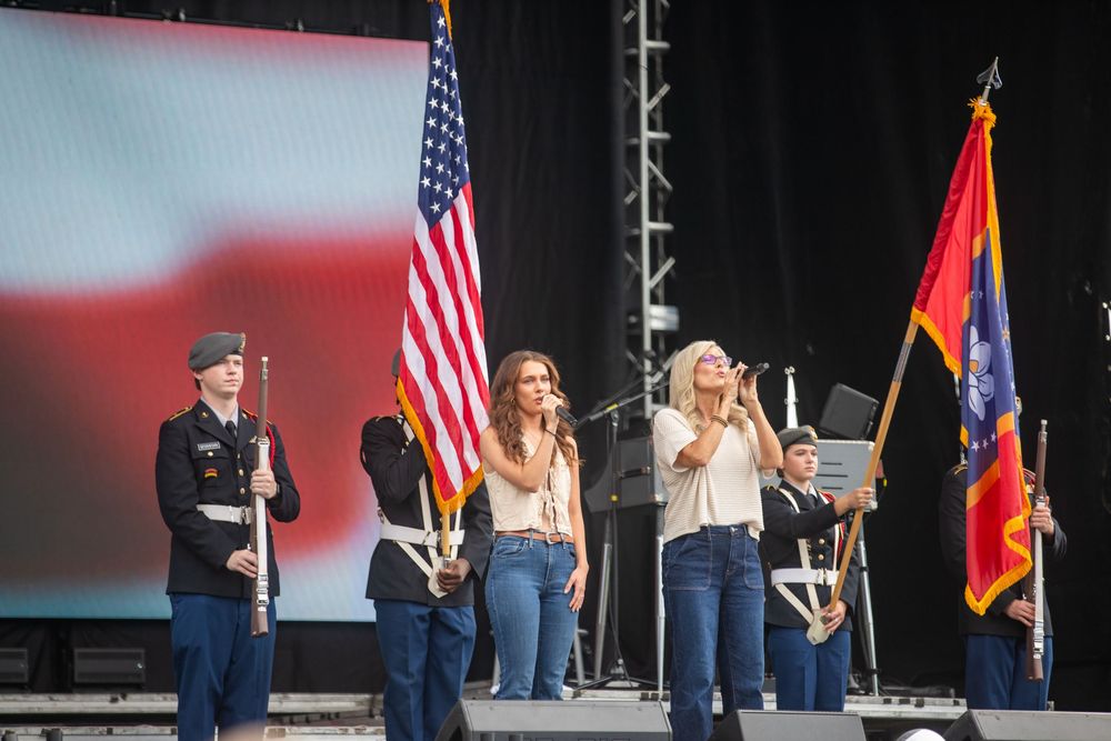 Two women perform on stage with military color guard holding US flag at an outdoor patriotic event.