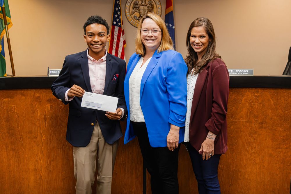 Three people pose for a photo holding a check at an official government building ceremony.